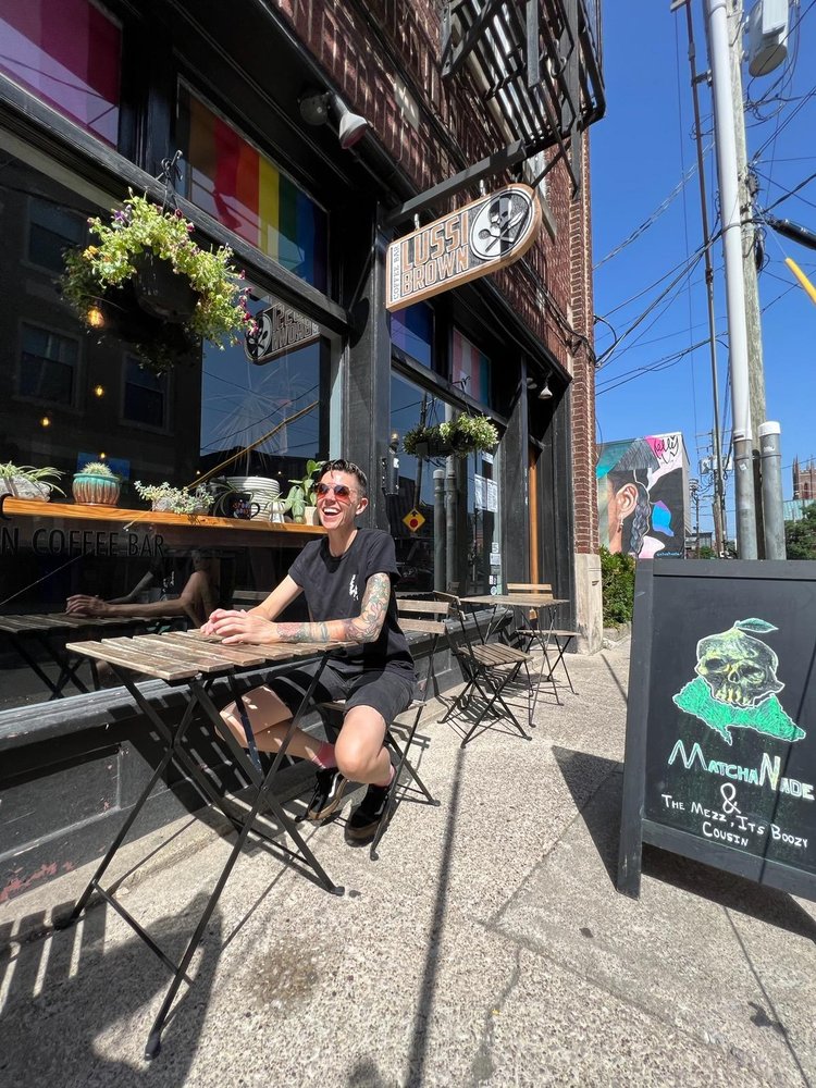 A person in a black shirt and shorts sits at a wooden table outside a coffee shop with a rainbow Pride flag hanging in the window